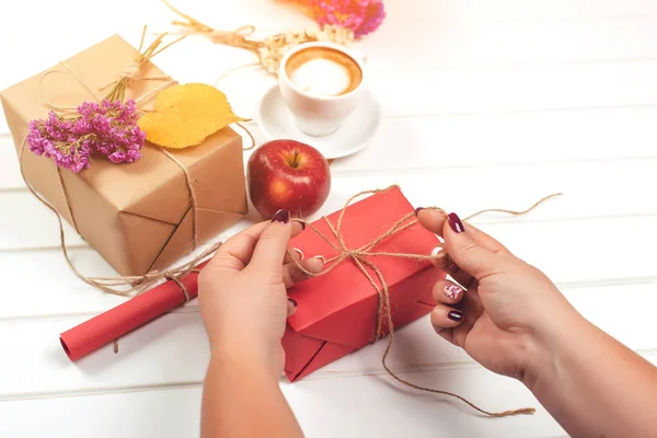 Woman preparing gifts for autumn holidays, birthday or thanksgiving day. Autumn composition. Handmade wrapped gift boxes, autumn leaves, apple, flowers, cup of coffee on white wooden background