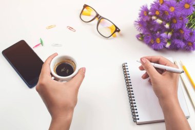 Woman's hands with coffee on office desk. Workplace with notebook, phone, glasses, pen, lilac flowers, cup of coffee. Flat lay, top view. Woman taking notes at workplace. Female workplace clipart