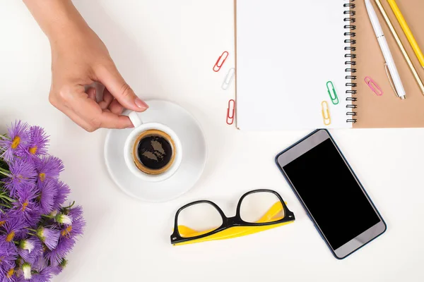Female workspace with cup of coffee, lilac flowers, eyeglasses and phone. Ideas, notes or plan writing concept. Top view. Flat lay. Woman hand hold cup of coffee. Woman working desk