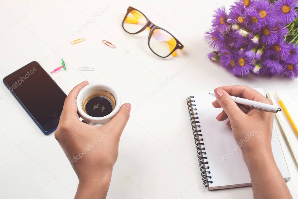 Woman's hands with coffee on office desk. Workplace with notebook, phone, glasses, pen, lilac flowers, cup of coffee. Flat lay, top view. Woman taking notes at workplace. Female workplace