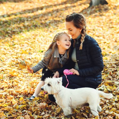 Mutlu bir aile ile köpek sonbahar parkta yürüyüş. Genç Anne ve kızı ile beyaz köpek düşen yapraklar eğleniyor. Sonbahar tatil. Aile, pet, Evcil hayvan ve insan kavramı. Mutlu çocukluk