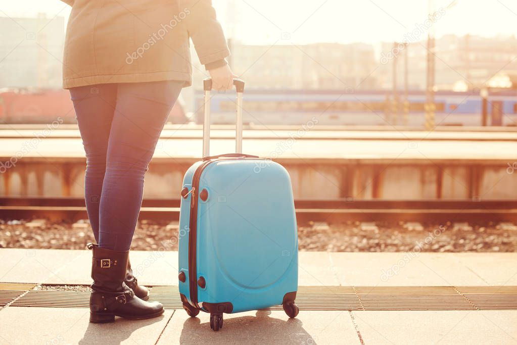 Traveler woman waiting at railway station. Travel by train. Girl standing with blue luggage suitcase in morning. Journey concept. Lifestyle, travelling, vacation. Autumn, winter holidays. Copy space