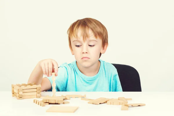 Sad child sitting at table. Child plays with construction toy blocks at the table. Sad bored boy build wooden house. Kid plays with wooden blocks. Sad child playing and dreaming about own home. Copy space