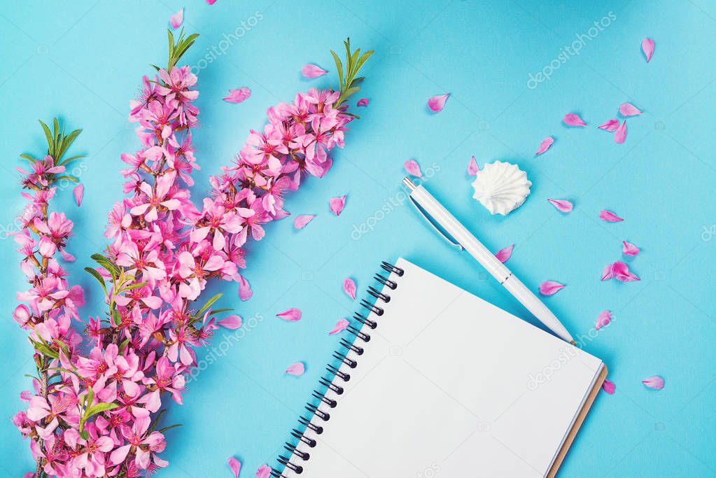 Opened notebook and spring flowers. Mock up. Spring pink flowers on blue paper background. Empty notebook for planning day, white pen and pink flowers. Woman working desk. Flat lay, top view