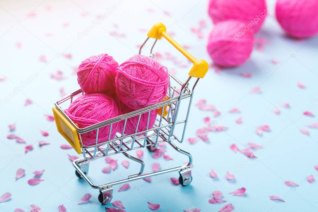 Shopping cart full of wool knitting balls. Knitting background. Pink wool yarns. Colorful pink threads on blue paper background. Seasonal sales. Spring shopping. Pink petals flowers on paper