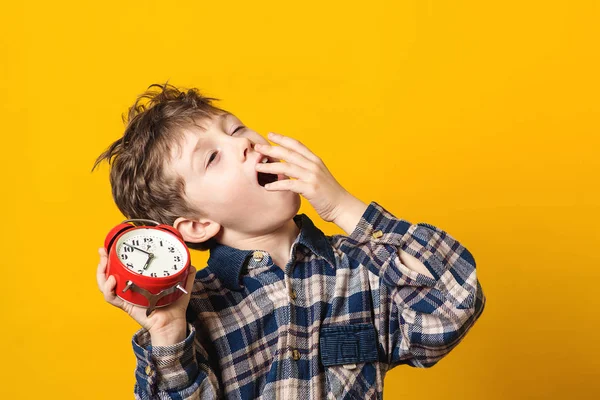 Sleepy and yawning schoolboy with alarm clock on yellow background. Funny boy yawns wide, covering his mouth with hand. Emotion concept. Cute yawning kid. Copy space