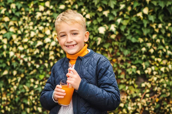 Happy cute boy drinking juice. Kid holding glass of orange juice at greenery background. Fresh orange juice outdoors. Healthy lifestyle. Happy childhood