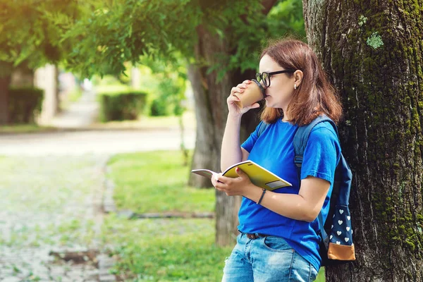 Jovem bebendo café e lendo um livro no parque de verão. Estudante menina gosta de recreação ao ar livre . — Fotografia de Stock