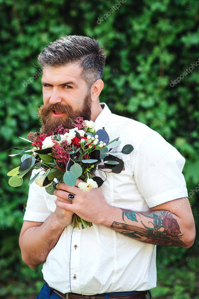 Excite bearded groom holds wedding bouquet. Handsome man with beautiful bouquet in his hands. Stylish bearded guy with a bouquet of red flowers and greens on greenery nature background. Wedding day.