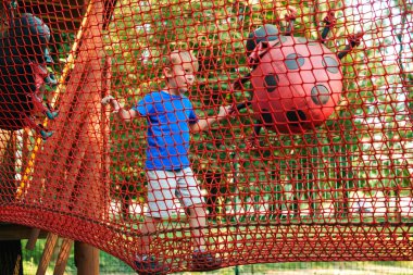Happy boy overcomes obstacles in rope adventure park. Summer holidays concept.  clipart