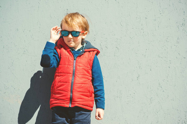 Stylish kid in trendy sunglasses. Kids fashion. Cute little blondy boy in red jacket standing over grey wall outdoors in sunny day. 