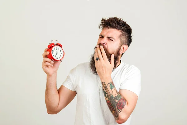 Hipster bostezando y sosteniendo el despertador. A las 7 de la mañana. El barbudo se levanta temprano por la mañana. . — Foto de Stock