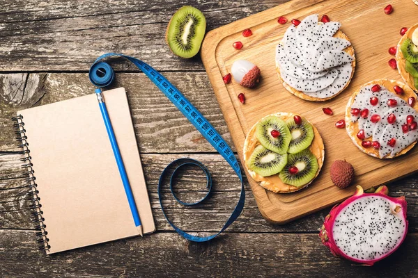 Planificación del concepto de dieta saludable. Entrenamiento y dieta fitness. Tostadas de frutas, cinta métrica y cuaderno vacío sobre fondo de madera . — Foto de Stock