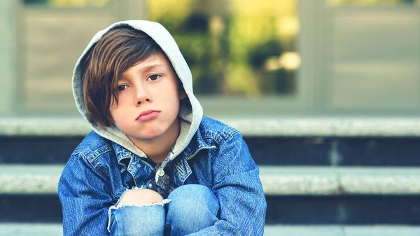 Trauriger Schuljunge, der draußen auf einer Treppe sitzt. unglücklicher Junge in Jeansjacke mit Kapuze und abgeschabtem Knie. — Stockfoto