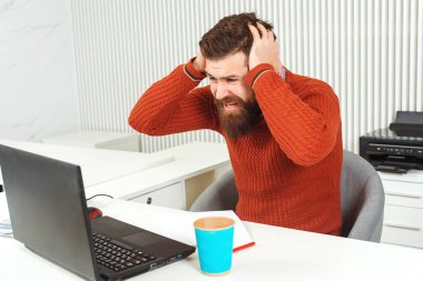 Stresli sakallı adam işyerinde eliyle başını tutarak. Dizüstü bilgisayara bakan adam.
