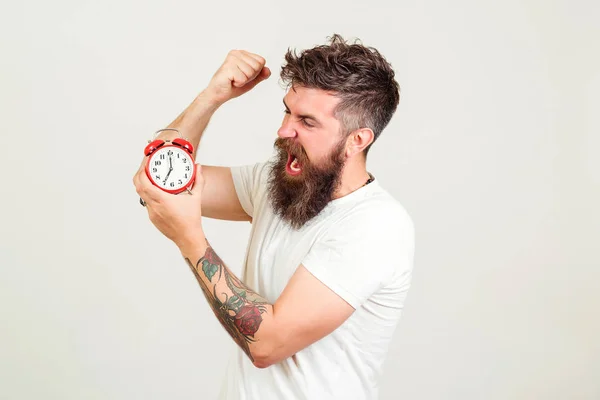El hombre barbudo enojado rompe el despertador. Hipsater brutal con tatuaje quiere destruir el reloj. Hora de trabajar . — Foto de Stock