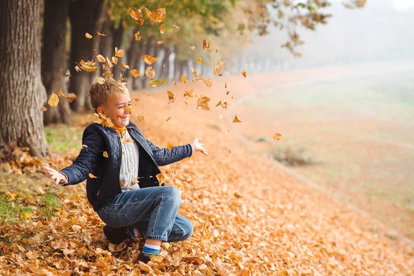 Glücklicher stilvoller Junge, der die gefallenen Blätter aufwirft. Nettes Kind beim Spaziergang an einem sonnigen Herbsttag. — Stockfoto