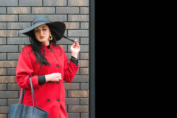 Mulher elegante com maquiagem perfeita, usando chapéu preto e casaco vermelho. Menina da moda posando perto de tijolo parede moderna do centro comercial, com espaço de cópia. Estilo de moda feminina — Fotografia de Stock