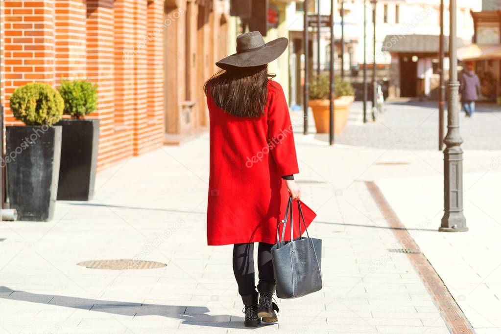 Stylish beautiful woman walking in street. Girl wearing red coat, black hat and holding trendy bag. Fashion outfit, autumn trend, accessories.