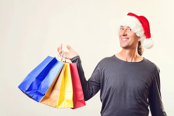 Noel Baba şapkalı yakışıklı adam alışveriş torbaları tutuyor. Noel hediyeleri zamanı. Yeni yıl ve Noel tatili. Mutlu alışveriş yapan adam bir sürü Noel hediyesi alıyor. — Stok fotoğraf