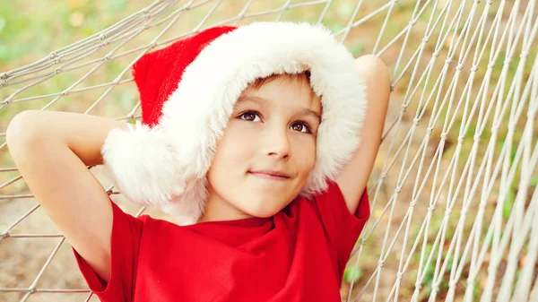 Cute little boy in Santa hat dreaming about christmas gifts. Christmas family vacation, travel, trip. Happy child lying in hammock in sunny day outdoors — Stock Photo, Image