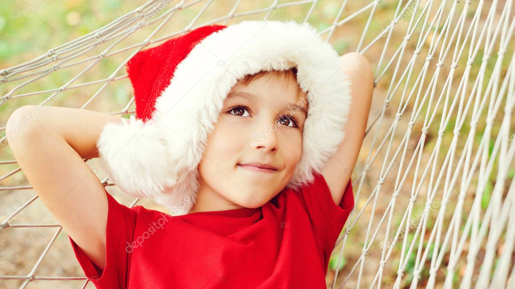 Cute little boy in Santa hat dreaming about christmas gifts. Christmas family vacation, travel, trip. Happy child lying in hammock in sunny day outdoors