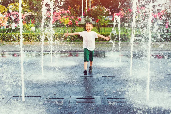 Happy boy running between water jets in city fountain. Summer in the city. Cute kid playing in a fountain with water. Child having fun in fountain. Summer vacation. Happy childhood.