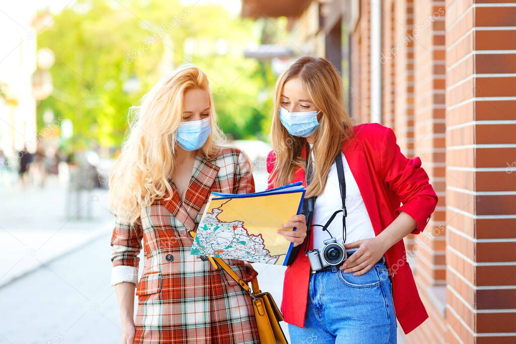 Stylish girls looking into tourist map in the city. Tourists girls are exploring new city together. Summer vacation during coronavirus pandemic. Women wearing face masks. Tourists in medical masked.