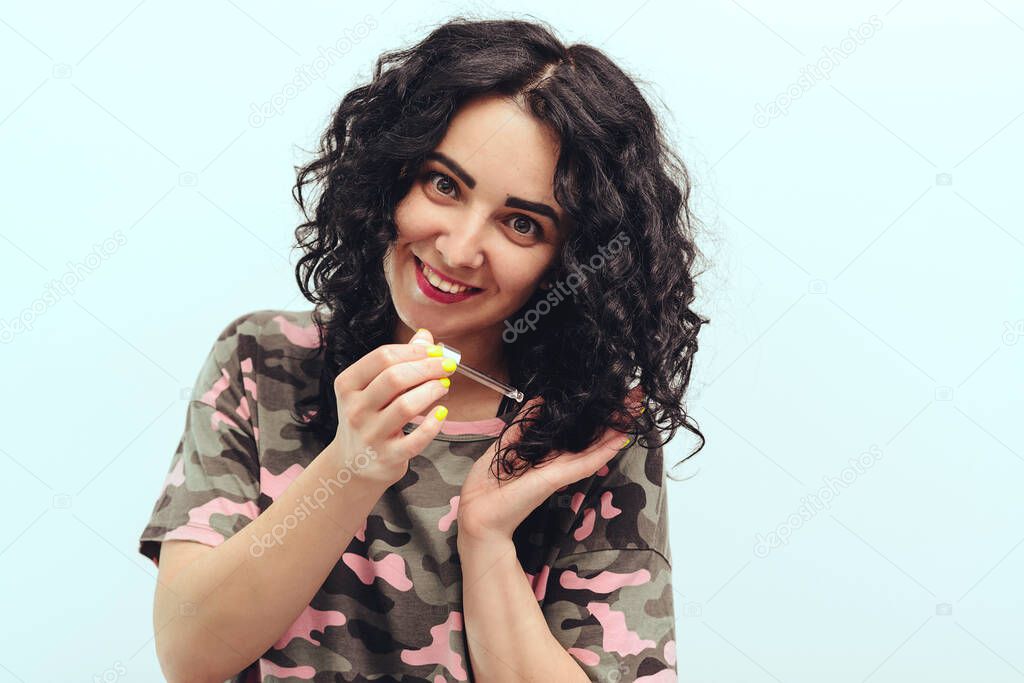 Happy girl applying natural oil on the tips of her curly hair at home. Natural oil hair treatment for woman. Curly woman using serum for hair. Products and cosmetics for hair.