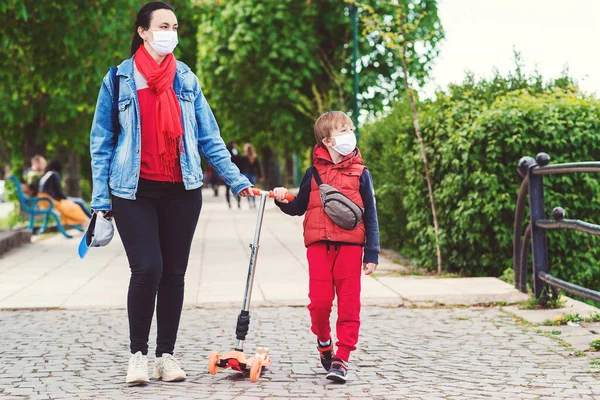 Family in safety masks outdoors. Riding boy on scooter in park. Boy wears medical face mask. Real life 2020. Coronavirus epidemic. Mother and son on a walk during coornavirus quarantine.
