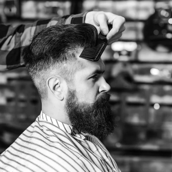 Bearded man getting haircut by hairdresser. Brutal guy sitting in barber chair. Vintage barbershop, business. Man making haircut to look perfect. Stylish bearded man in salon. New perfect style.