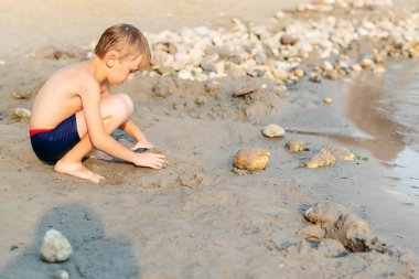 Kumsalda oyun oynayan tatlı bir çocuk. Küçük çocuk yaz plajında kumla oynuyor. Yaz tatilinde komik oyunlar. Çocuk sahilde kumla eğleniyor. Güneşli bir yaz günü. Boş zaman, oyunlar ve tatil.