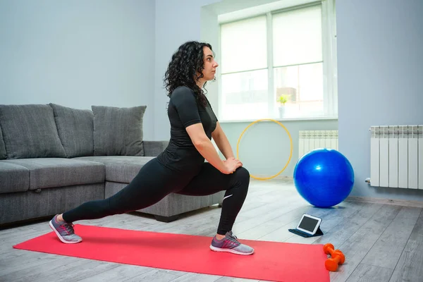 Woman stretching her leg and back. Online sport fitness yoga training. Young woman doing exercises on yoga mat. Girl using tablet looks online master class. Training at home.