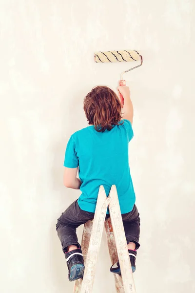 Young boy painting interior wall with paint roller. New house for family. Home renovation. Kid using paint roller. Son helps parents to paint wall. Boy sitting on the ladder.