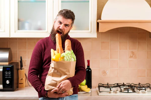 Hungry man bites baguette at kitchen. Bearded man holding paper bag with food at home. Delivery food, products to home. Shopping and healthy food. Online shop with food, products and drinks.