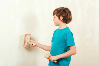 Şirin çocuk fırçayla bir duvar boyuyor. Fırçayı beyaz renge boyayan çocuk. İnşaat ve yenileme. Ev yenileme servisi. Odada duvar boyayan çocuk..