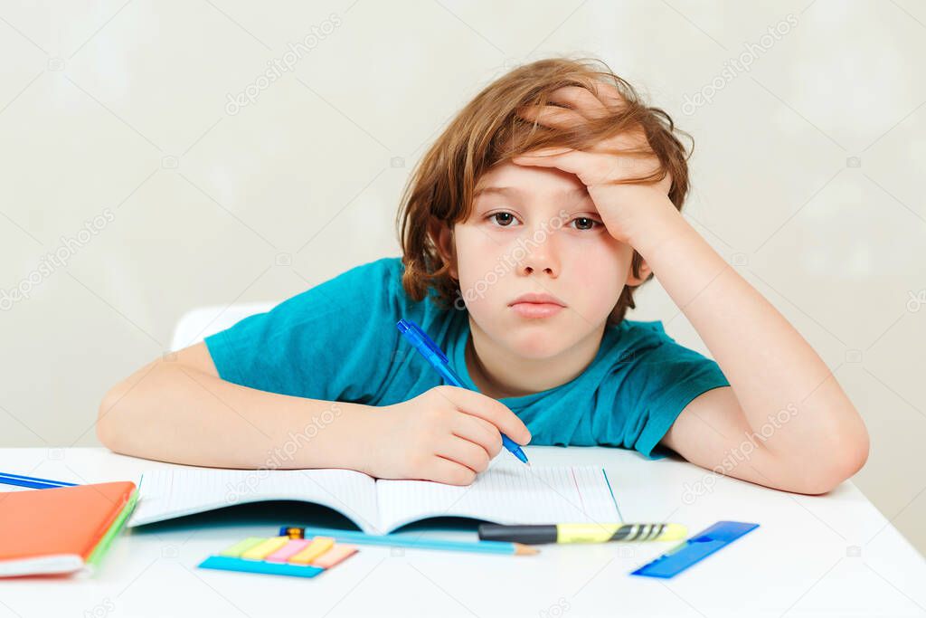 Tired schoolboy sitting at table. Boy doing homework. Learning difficulties, education concept. Stressed out and tired kid. Frustrated kid sitting at the desk with many books. Sad tired schoolchild.
