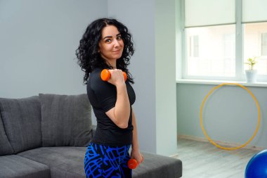 Spor, spor ve sağlıklı yaşam tarzı konsepti. Evde kız eğitimi. Kadınlar formda kalmak ister. Odada egzersiz yapan genç bir kadın. Kadın evde dambıllarla çalışıyor..