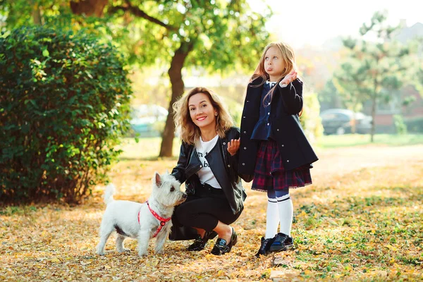 Happy family playing with dog in autumn park. Mother and daughter having fun together on a walk. Fashionable mother and schoolgirl walking with their dog in park. Autumn time, weekend and holidays.