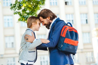 Baba çocuğu okula götürür. Okul konseptine geri dönelim. Öğrenci birinci sınıfa git. Eğitim, ilkokul. Baba, okula başladığı için oğluna destek oluyor. Sakallı adam mutlu öğrenciyi kucaklıyor..