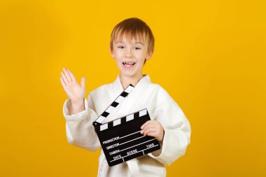 Beyaz kimonolu mutlu çocuk video ya da film çekiyor. Sarı arka planda küçük bir aktör. Çocuklar spor, hobiler ve eğlence konsepti. Çocuk elinde bir el çırpma tahtası tutuyor. Pozitif duygular ve ifade.