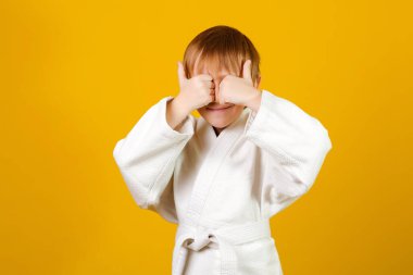 Beyaz kimono giyen bir çocuk. Mutlu karateci çocuk. Sağlıklı bir çocukluk. Dövüş sanatları okulu. Sağlıklı yaşam tarzı ve spor. Sarı arka planda beyaz kimonolu küçük bir çocuk. Başparmak gösteren çocuk.