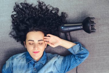 Beauty woman with curly hair lying on sofa. Girl styling her hair with modern hairdryer. Professional hairdryer with special diffuser nozzle. Woman drying hair at home. Fashion and beauty. clipart
