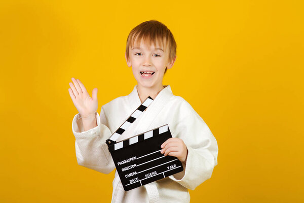Happy child in white kimono making video or movie. Little actor over yellow background. Kids sport, hobbies and leisure concept. Boy holding a film clapper board. Positive emotions and expresion.