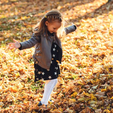 Mutlu kız çocuğu sonbahar yapraklarını dışarı fırlatıyor. Sonbahar parkında şık deri ceketli ve elbiseli küçük sarışın kız. Mutlu çocukluk, sezon ve insanların konsepti. Sonbahar çocuk modası