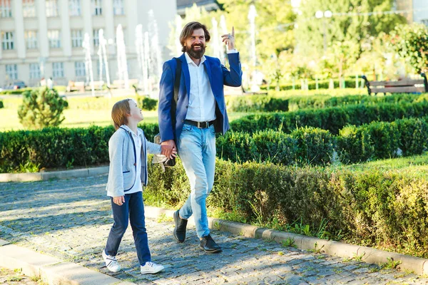 Father and son in suits walking at city street. Stylish dad and kid go hand in hand. Father talking with son outdoors. Happy family going home after school. Fashion, fatherhood and relationship.