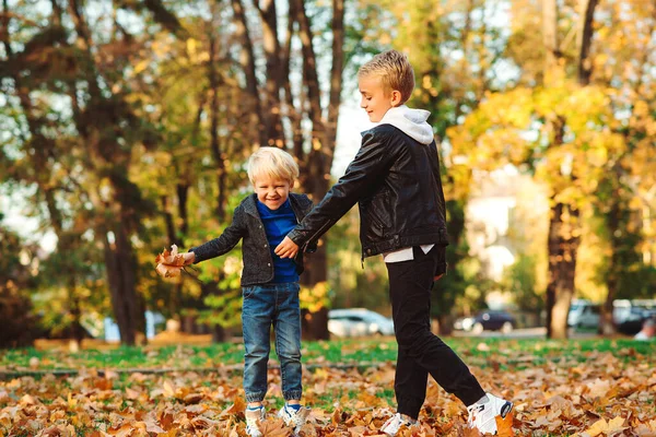 Happy brothers playing together in autumn park. Stylish children throwing autumn leaves. Kids on a walk in autumn day. Happy and healthy childhood. Family, fashion, vacation and autumn concept