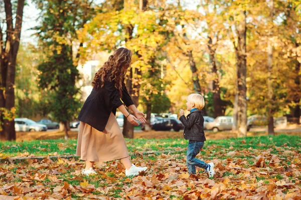 妈妈和孩子一起在秋天的公园里玩耍 秋天散步时 快乐的年轻家庭 妈妈和小儿子在享受秋天 母性快乐秋天的时尚 用落叶寻欢作乐的时髦家庭 — 图库照片