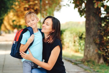 Sırt çantalı akıllı çocuk parkta annesine sarılıyor. Okul konseptine geri dönelim. Mutlu çocuk ve anne aynı okula gidiyor. Okulun ilk günü..