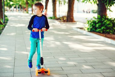 Yaz parkında scooter süren sevimli bir çocuk. Sağlıklı yaşam tarzı ve spor eğlencesi. Çocuk dışarıda aktif zaman geçiriyor. Çocuklar paten kaymayı öğreniyor. Mutlu çocukluk.